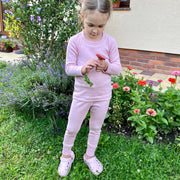 Young girl in a pink merino wool thermal base layer set standing in a garden with flowers and plants.