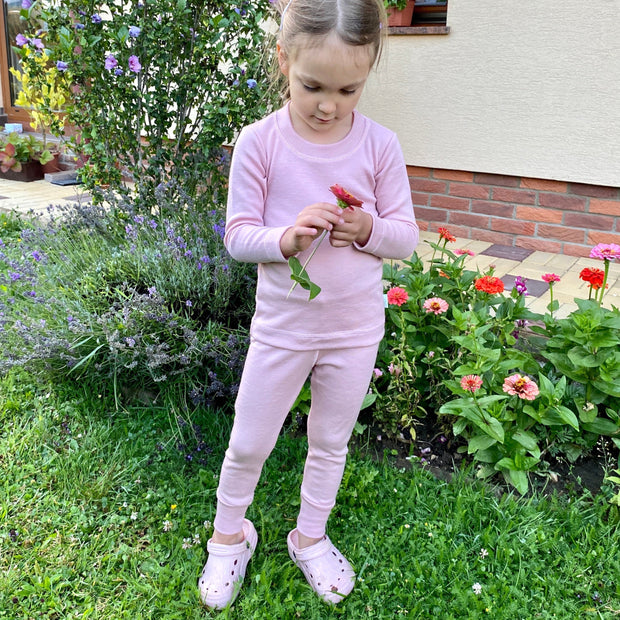 Young girl in a pink merino wool thermal base layer set standing in a garden with flowers and plants.