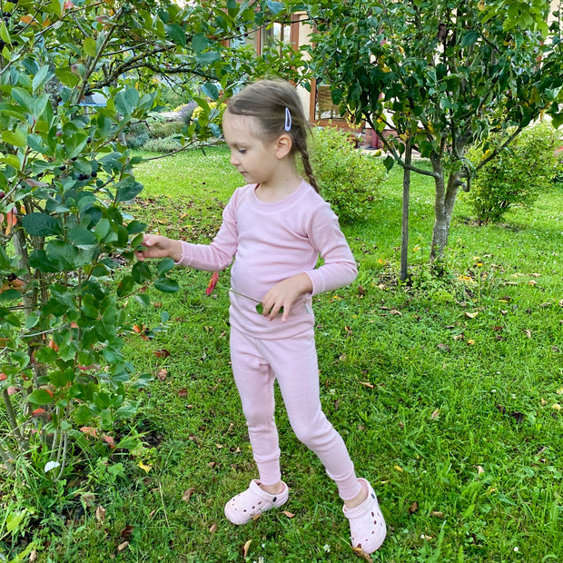 Young girl in a pink merino wool base layer set standing in an orchard among green trees.